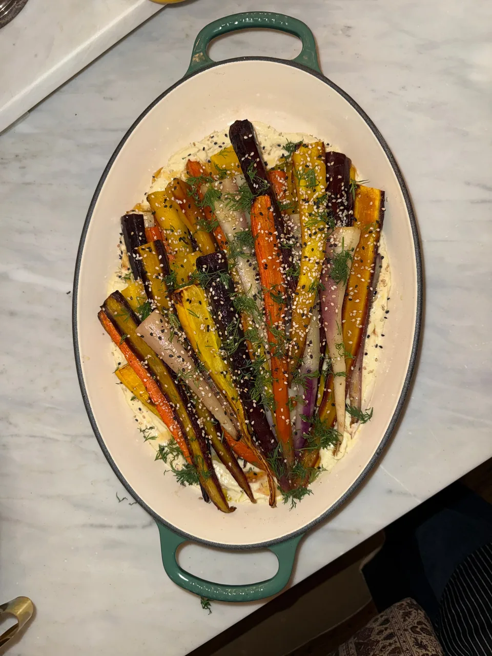 Roasted rainbow carrots over a creamy spread, topped with dill and sesame seeds, in a green enameled dish