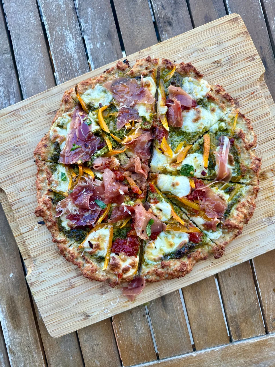 Pizza with pesto, melted mozzarella, prosciutto and sliced yellow peppers on a wood board